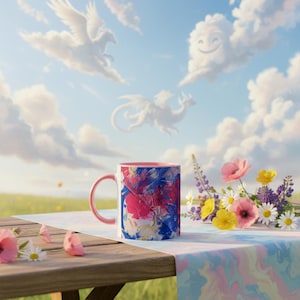 May include: A ceramic mug with a pink handle and interior, featuring an abstract design in shades of blue, pink, and white. The mug sits on a wooden table with a floral arrangement and a pastel tablecloth. The background shows a whimsical sky with cloud formations.