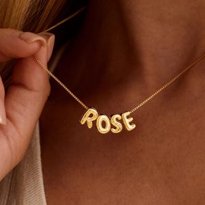 May include: A gold necklace spelling out the name "ROSE" in capital letters. The necklace is worn around the neck. The image focuses on the necklace and the name. The background is blurred.