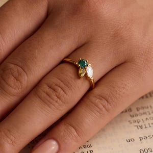 Könnte beinhalten: Ein goldener Ring mit einem Cluster aus Edelsteinen. Der Ring hat einen zentralen smaragdgrünen Stein, flankiert von zwei kleineren, birnenförmigen Steinen, einem weißen und einem gelben. Der Ring wird an einer Hand gezeigt.