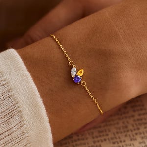 May include: A delicate gold chain bracelet featuring three small, faceted gemstones arranged in a cluster. The stones include a clear marquise-cut gem, a yellow pear-cut gem, and a round purple gem, all set in a simple gold metal frame.