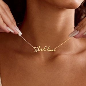 May include: A gold-coloured necklace featuring a delicate chain and a central nameplate pendant in a cursive script font that reads the name Stella. The metal has a polished finish and rests against the neck, held by hands on both sides.