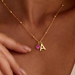 Peut inclure: Un collier doré avec un pendentif en forme de lettre "A" et une pierre précieuse rose. La chaîne est délicate, avec de petites perles espacées. Le pendentif est suspendu près du centre de la poitrine.