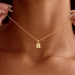 May include: A gold-coloured necklace featuring a pendant in the shape of the letter "R". The necklace has a delicate, thin chain. The pendant is a capital letter "R" in a rounded font.