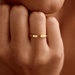 May include: A gold-toned open ring featuring two flat, rounded ends. Each end is engraved with a single uppercase letter, specifically the letter M on the left side and the letter A on the right side, creating a personalised initial design.