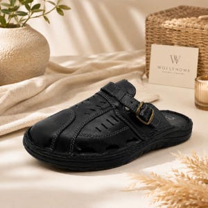 Könnte beinhalten: Ein schwarzer Pantoletten-Schuh aus Leder mit geschlossener, abgerundeter Zehenpartie, Ziernähten und seitlichen Belüftungsschlitzen. Der Schuh verfügt über einen verstellbaren Riemen am Spann mit einer bronzefarbenen Metallschnalle.