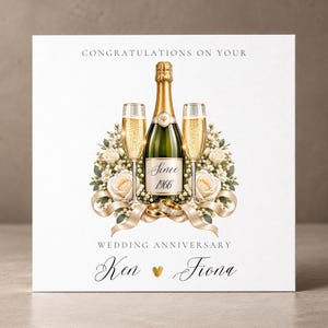 Könnte beinhalten: Eine quadratische Grußkarte mit einer Champagnerflasche und zwei Sektgläsern, umgeben von weißen Rosen und Bändern. Der Text lautet CONGRATULATIONS ON YOUR WEDDING ANNIVERSARY, Since 1966, sowie Ken und Fiona mit einem goldenen Herz. Zwei goldene Ringe.