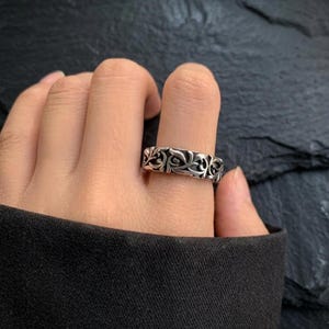 May include: Silver-colored ring with an ornate, repeating floral design. The ring is worn on a finger, showcasing the detailed pattern of swirling leaves and curves. The ring's metal has a slightly aged appearance.