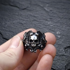 Peut inclure: Une bague argentée ornée d'un crâne. Le crâne est le point central, avec des traits faciaux détaillés. La bague présente un motif orné de style gothique autour du crâne.