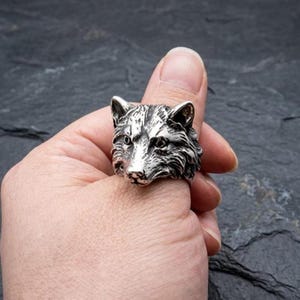 Puede incluir: Anillo plateado con un diseño detallado de cabeza de lobo. El anillo muestra los rasgos faciales del lobo, incluyendo sus ojos, nariz y textura del pelaje. El anillo se lleva en un dedo, resaltando la intrincada artesanía.