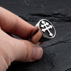 Könnte beinhalten: Ein silberner Ring mit einem Doppelkreuz-Design. Der Ring hat eine polierte Oberfläche und einen schwarzen Hintergrund. Das Kreuz ist weiß und hat ein detailliertes Design. Der Ring wird von einer Hand gehalten.
