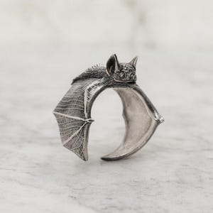 Puede incluir: Un anillo de murciélago plateado. El anillo presenta un diseño detallado de murciélago con alas extendidas. La cabeza del murciélago está en la parte superior del anillo, y las alas se curvan alrededor del dedo. El anillo es de metal brillante.