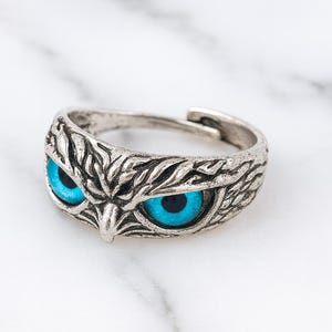 Könnte beinhalten: Ein verstellbarer Ring in Silberoptik mit einem Eulen-Design. Der Ring zeigt das Gesicht der Eule mit detaillierten Federstrukturen und leuchtend blauen Augen. Der Ring ist vor einem weißen Marmorhintergrund platziert.
