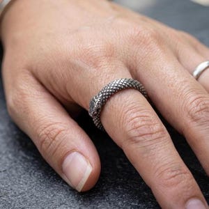 May include: A silver snake ring, coiled around a finger. The ring features a textured, scale-like design and a detailed snake head. The ring is worn on a finger, showcasing its intricate design.