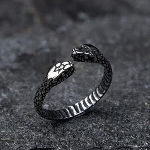 May include: A silver-coloured ring in the shape of a snake with two heads. The ring has a textured, scale-like pattern on the band and snake bodies. The snake heads face each other, creating an open design.