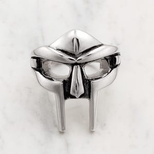 Puede incluir: Un colgante plateado con forma de máscara. La máscara tiene un diseño distintivo con una parte superior puntiaguda, aberturas rectangulares para los ojos y una nariz prominente. El colgante es probablemente de metal y tiene un acabado pulido.