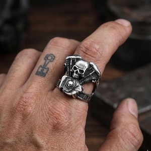 May include: A silver-colored ring featuring a skull and engine design. The ring has intricate details, including the skull's facial features and engine components. The ring is worn on a finger, showcasing the biker-themed jewelry.