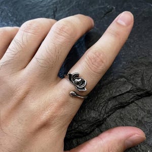 May include: A silver-coloured ring featuring a lotus flower design. The ring has an adjustable band and the lotus flower is detailed with petals and a central bud. The ring is worn on a finger.