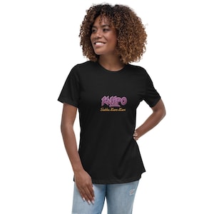 Peut inclure: T-shirt noir avec le mot "Kalipo" en violet, dans un style graffiti. En dessous, le texte "Sakku Kam-Kam" est affiché. Le t-shirt est de coupe décontractée.