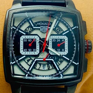 May include: A black TAG Heuer Monaco watch with a square case and a black metal link bracelet. The dial features a skeletonized design, two white square sub-dials, a date window at the bottom, red hands, and the text Monaco TAG Heuer Swiss Made.
