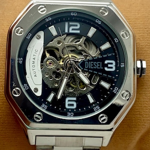 May include: A silver-toned stainless steel automatic wristwatch featuring an octagonal bezel with exposed screws, a dark blue skeleton dial revealing internal gears, white hour markers, and the Diesel logo printed at the three o'clock position.