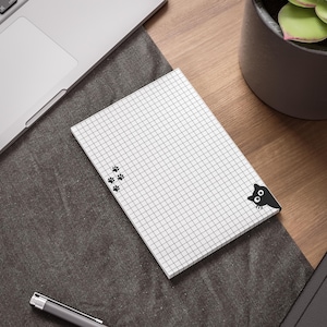 May include: A rectangular notepad with a white grid pattern on a light background. The notepad features a small black cat illustration peeking from the bottom right corner and a trail of four black paw prints on the left side of the grid.