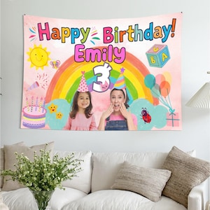May include: A rectangular birthday wall tapestry featuring a pink background with a large rainbow, a yellow sun, a birthday cake with candles, a ladybug, and a blue alphabet block. The text reads Happy Birthday! Emily with a large number 3.