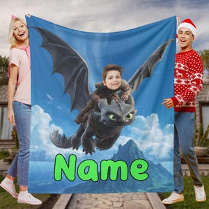 Könnte beinhalten: Eine blaue Decke mit einem Zeichentrickdrachen und einem Kind, das vor einem blauen Himmel mit Wolken und Bergen reitet. Das Wort "Name" steht grün am unteren Rand. Die Decke wird von zwei Personen gehalten.
