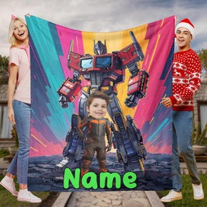 Puede incluir: Una colorida manta con una gran ilustración de Optimus Prime y la cara de un niño. La manta tiene un fondo vibrante con la palabra "Name" en verde en la parte inferior. La manta está sujeta por dos personas.