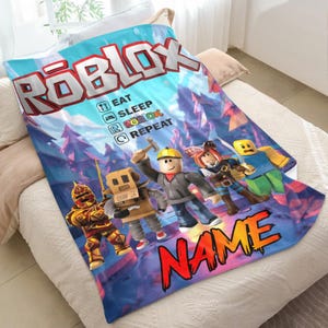 May include: A blue and white Roblox blanket with the Roblox logo and characters. The blanket features the text "EAT SLEEP ROBLOX REPEAT" and a space for a name. The background depicts a forest scene.