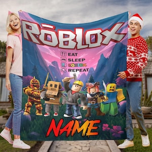 May include: A Roblox blanket with the Roblox logo, the text "EAT SLEEP ROBLOX REPEAT," and Roblox character illustrations. The blanket has a colourful design with a mountain and sky background, and the word "NAME" in large, red letters.