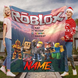 May include: A rectangular Roblox blanket with the Roblox logo and character illustrations. The blanket has the text "EAT SLEEP ROBLOX REPEAT" and a name at the bottom. The background depicts a sunset. The blanket is likely made of soft fabric.