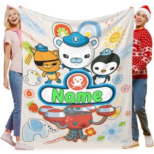 Puede incluir: Una manta de forro polar blanca personalizada con personajes de dibujos animados, incluyendo un oso, un pingüino y un gato, con la palabra "Name" en un diseño azul y verde. La manta tiene un vehículo rojo y naranja con forma de submarino en la parte inferior.