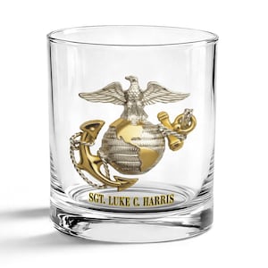 May include: A clear glass tumbler with a detailed emblem featuring an eagle, globe, and anchor in gold and silver tones. The text "SGT. LUKE C. HARRIS" is printed below the emblem. The glass is cylindrical and sits on a white surface.