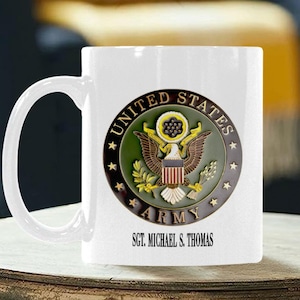 Könnte beinhalten: Weiße Keramiktasse mit dem Emblem der United States Army, das einen Adler, Olivenzweige und Sterne zeigt. Der Text "UNITED STATES ARMY" und "SGT. MICHAEL S. THOMAS" ist auf der Tasse aufgedruckt. Die Tasse steht auf einem Holztisch.