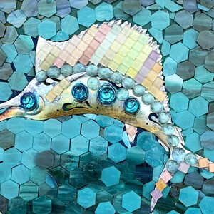 Puede incluir: Una obra de arte de mosaico redondo con un pez vela. El pez está detallado con colores iridiscentes y detalles de vidrio azul. El fondo está compuesto por baldosas hexagonales turquesas y verde azulado, enmarcadas por un borde de madera clara.
