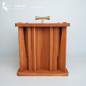 Puede incluir: Un mueble de madera con un acabado marrón cálido. El mueble presenta listones de madera verticales y un asa dorada. La parte superior del mueble tiene una serie de pequeñas clavijas de madera. El texto "fofo lab forming the formless" y "PLUZ series" son visibles.