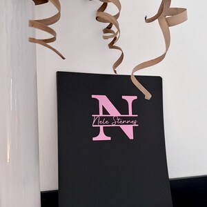 Könnte beinhalten: Ein schwarzes Notizbuch mit einem rosa Monogramm des Buchstabens "N" und dem Namen "Nele Stennes". Das Notizbuch liegt auf einem schwarzen Regal, mit einer weißen Vase und braunen Papierlocken im Hintergrund. Thema: Schreibwaren.