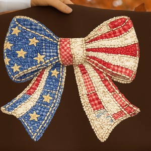 May include: A decorative bow in patriotic colours against a brown backdrop. The bow is made of blue fabric with gold stars and red and white striped fabric. The bow appears handmade and has a stitched design.