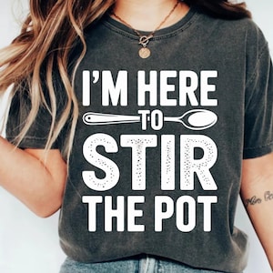 Może przedstawiać: Ciemnoszara koszulka z białym napisem "I'M HERE TO STIR THE POT". Projekt zawiera grafikę łyżki. Koszulka jest w stylu casual.