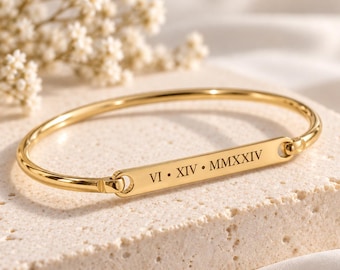 May include: A gold-toned metal bangle bracelet featuring a central horizontal bar engraved with the Roman numerals VI • XIV • MMXXIV. The bracelet has a polished finish and a hinged design, showcasing a clean, metallic aesthetic with clear lettering.