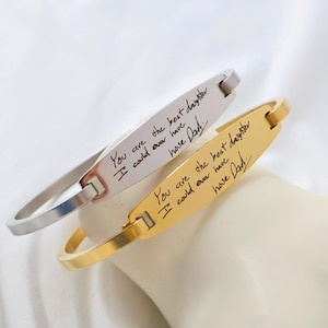 Könnte beinhalten: Zwei gravierte Armbänder, eines silberfarben und eines goldfarben, jeweils mit der handschriftlichen Inschrift "You are the best daughter I could ever have. Dad." Die Armbänder werden auf einer weißen Oberfläche präsentiert.