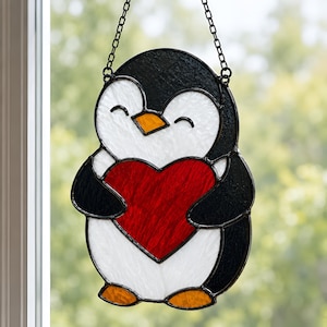 Könnte beinhalten: Ein Buntglas-Suncatcher in Form eines Pinguins mit schwarzen, weißen und orangefarbenen Glasteilen. Der Pinguin hält ein großes, strukturiertes rotes Herz in der Mitte. Das Stück ist mit dunklem Metalllot eingefasst und hängt an einer schwarzen Kette.