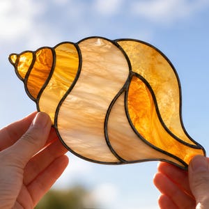 Könnte beinhalten: Ein Sonnenfänger aus Buntglas in Form einer Muschel, bestehend aus durchscheinenden bernsteinfarbenen und cremefarbenen Glassegmenten, die durch dunkle Metalllötlinien verbunden sind und eine Spiralform bilden, durch die das Licht scheint.