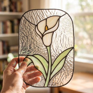 May include: A rectangular stained glass panel featuring a central white calla lily with a yellow spadix and light green leaves. The flower is set against a clear, textured glass background, all held together by dark metal solder lines.