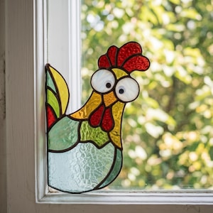 May include: A stained glass suncatcher shaped like a chicken with a red comb, yellow head, and a body featuring textured light blue, green, and yellow glass panels. The chicken has two large white circular eyes with black pupils.