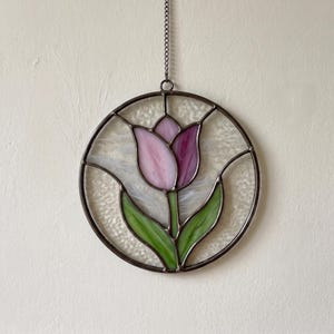 May include: A circular stained glass suncatcher featuring a pink and purple tulip flower with green leaves. The design is encased in a dark metal frame with a textured white background and includes a metal chain for hanging purposes.