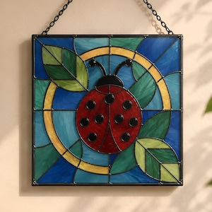 May include: Stained glass panel depicting a red ladybird with black spots, set against a blue and green backdrop. The ladybird is surrounded by green leaves and a yellow circle. The panel is framed in black and hangs from a chain.