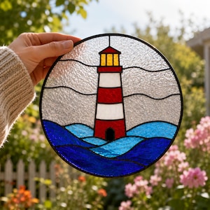 May include: Round stained glass art piece featuring a lighthouse with red and white stripes, a yellow light, and blue waves. The lighthouse is set against a textured, clear glass background, framed by a black border. A nautical-themed decorative item.