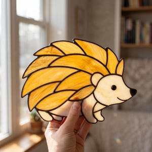 May include: A stained glass hedgehog suncatcher featuring a body made of textured amber and yellow glass pieces with black solder lines. The head and underbelly are crafted from smooth, opaque cream-colored glass with a small black glass eye.