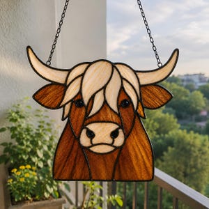 May include: A stained glass suncatcher shaped like a Highland cow head, featuring textured brown and cream glass pieces held together by black metal solder lines. The piece includes two curved horns and a hanging chain attached to the top.